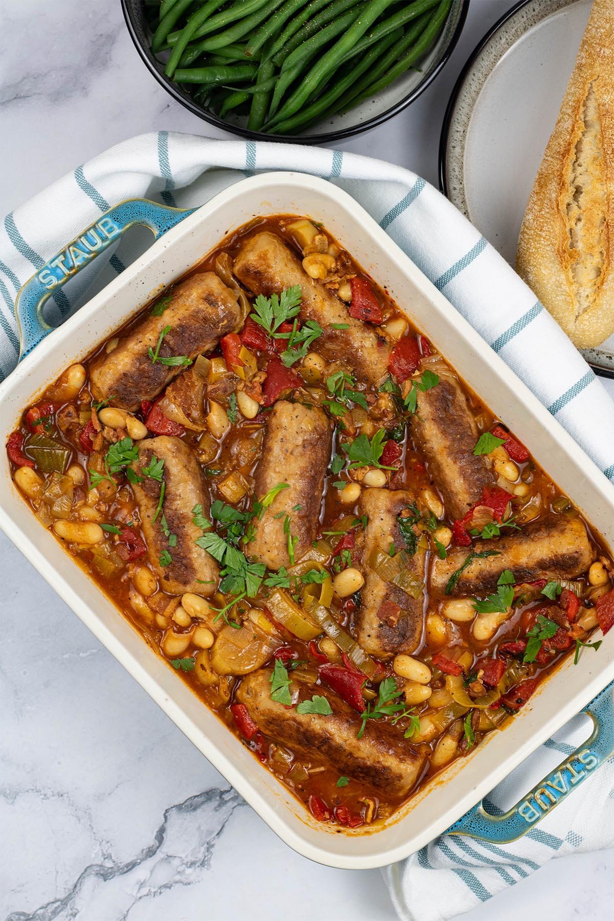 Slow cooker sausage casserole in a rectangle Staub serving dish with baguette and green beans in the background.