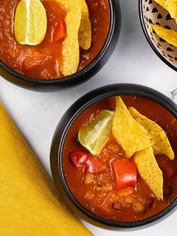 Two portions of spicy chilli bean soup in black bowls garnished with tortilla crisps and a wedge of lime.