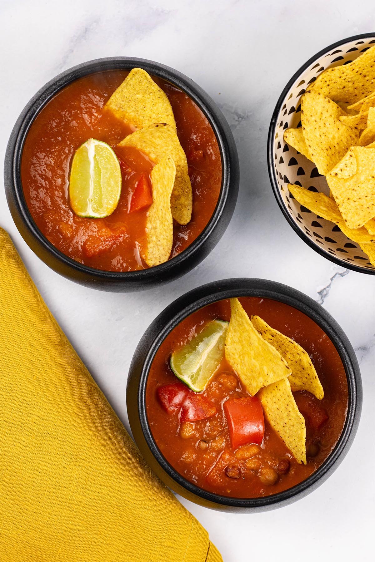 Two portions of spicy chilli bean soup in black bowls garnished with tortilla crisps and a wedge of lime.