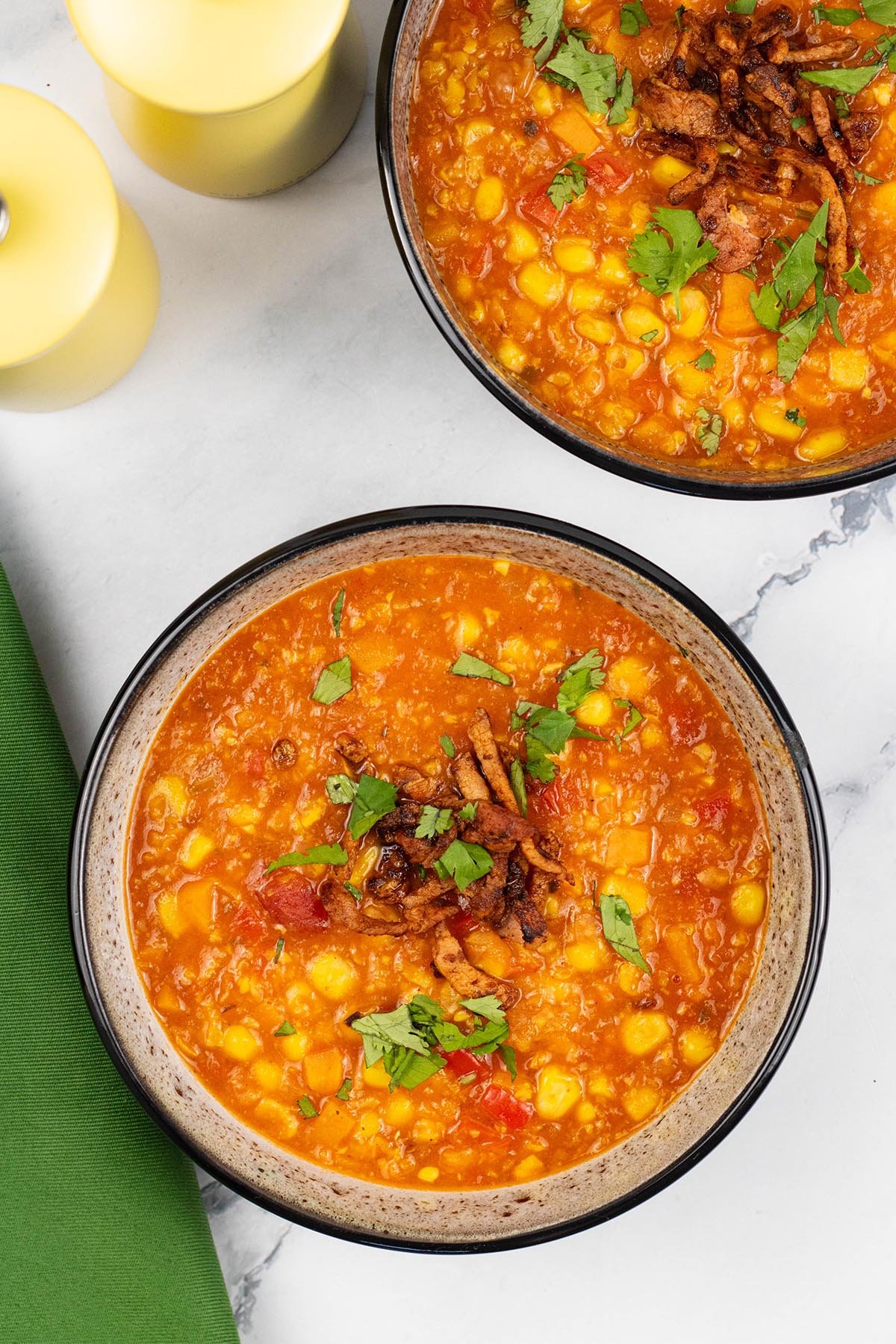 Two portions of spicy sweetcorn soup in black patterned bowls with yellow salt and pepper grinders and a green napkin on the side.