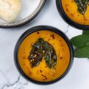 Two portions of squash and carrot soup with crispy sage leaves in black bowls with a plate of crusty rolls on the side.