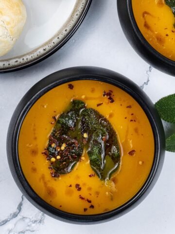 Two portions of squash and carrot soup with crispy sage leaves in black bowls with a plate of crusty rolls on the side.