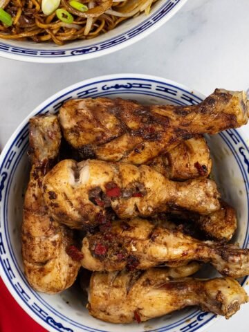 Sticky chicken drumsticks in a blue and white oriental serving bowl with a bowl of fried noodles with beansprouts in the background.